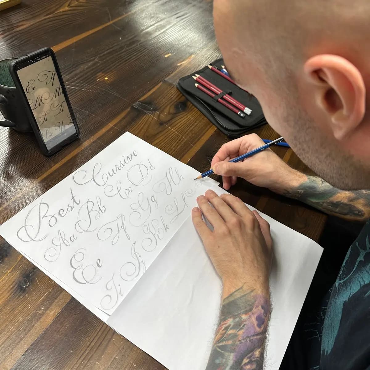 Person am Studiotisch zeichnet ein Alphabet auf Papier, daneben eine Vorlage am Handy